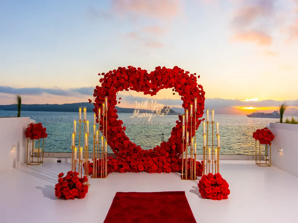 A red-rose heart arch on a Santorini cliff terrace overlooking the caldera and the Aegean Sea at golden hour