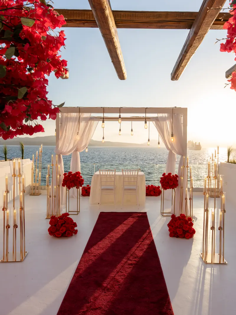 A meticulously styled proposal setup — a red carpet aisle lined with roses and candles leading to a draped arch — on a Santorini caldera terrace