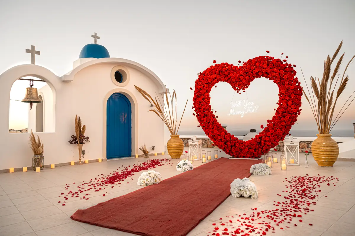 A white Cycladic chapel beside a red-rose heart arch and red carpet aisle overlooking the Santorini sea