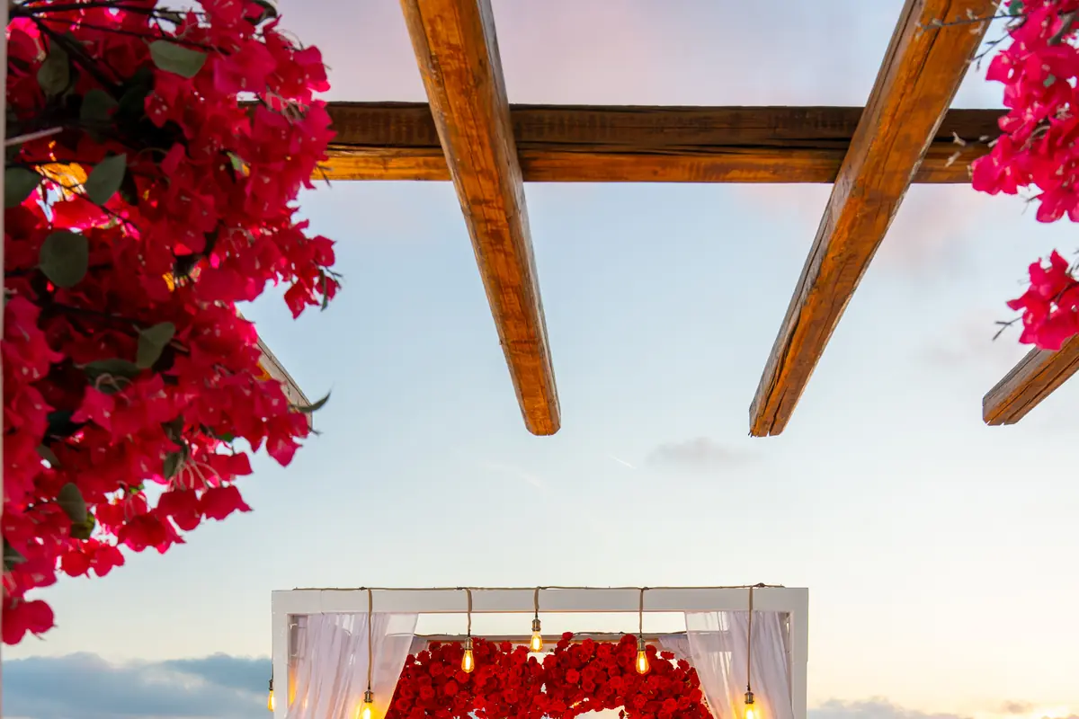 A red-rose heart arch framed by pink bougainvillea with a red carpet aisle on a Santorini terrace