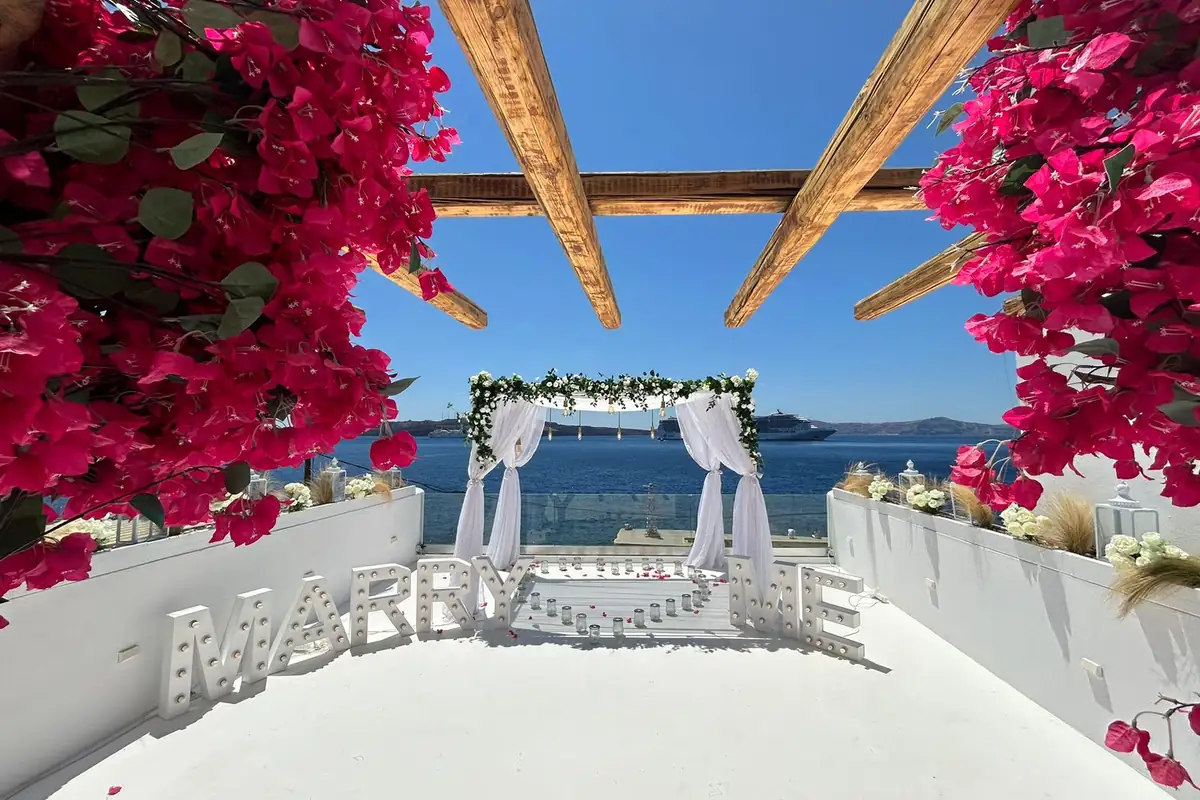 Vivid pink bougainvillea framing a MARRY ME proposal setup above the blue Santorini caldera