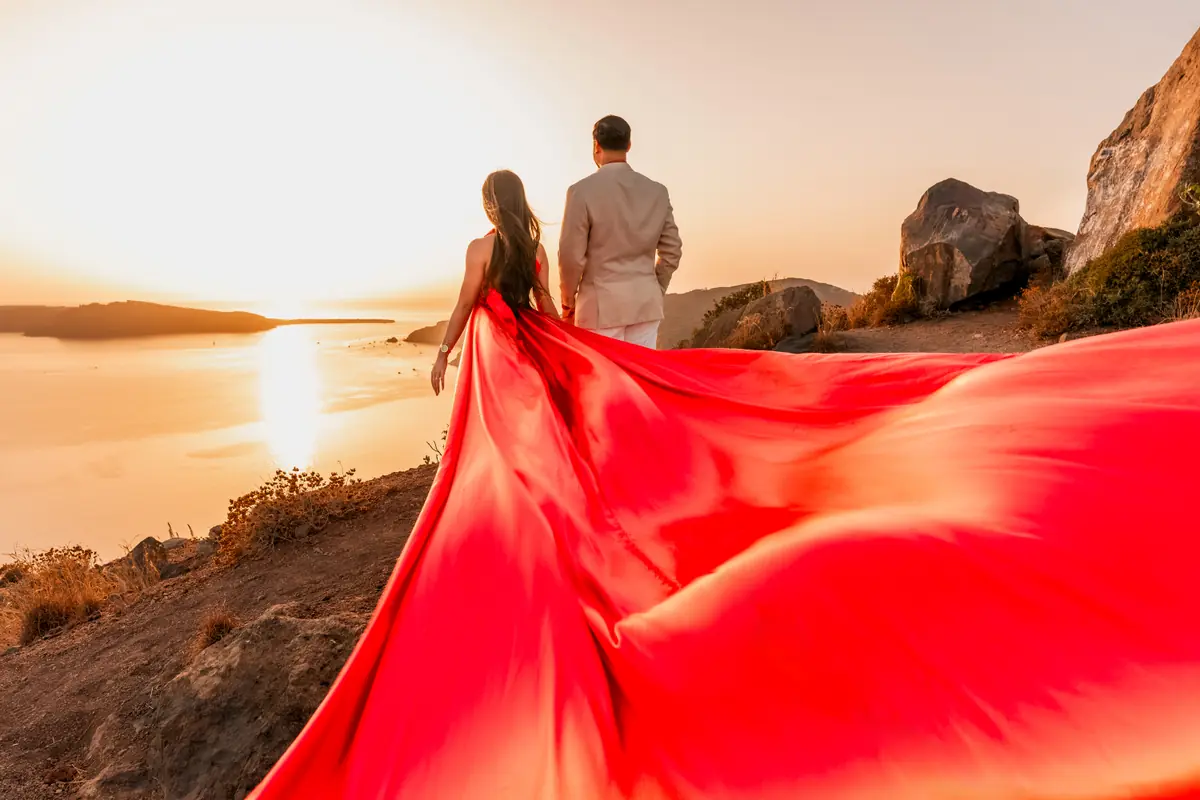 A couple with a dramatic flowing red dress on a Santorini clifftop as the sun sets over the sea