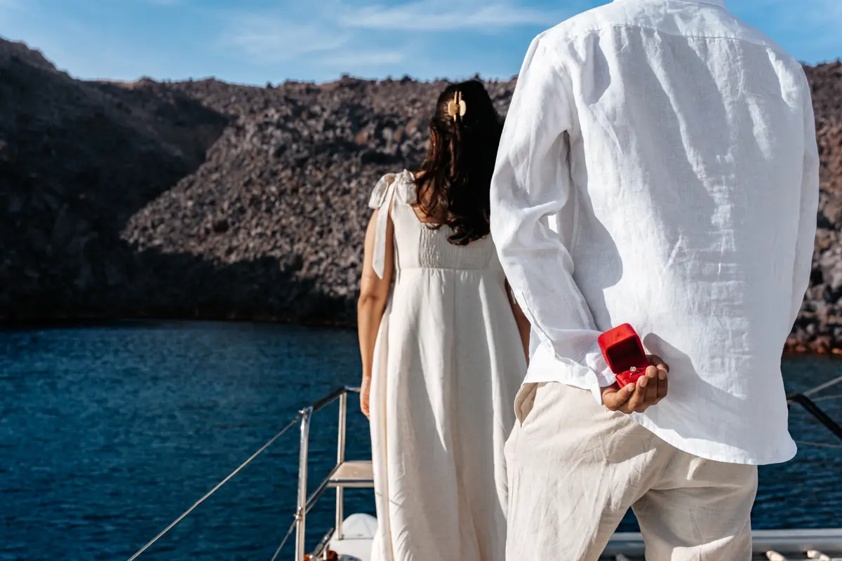 A man hides a red ring box behind his back beside his unsuspecting partner on a yacht off Santorini