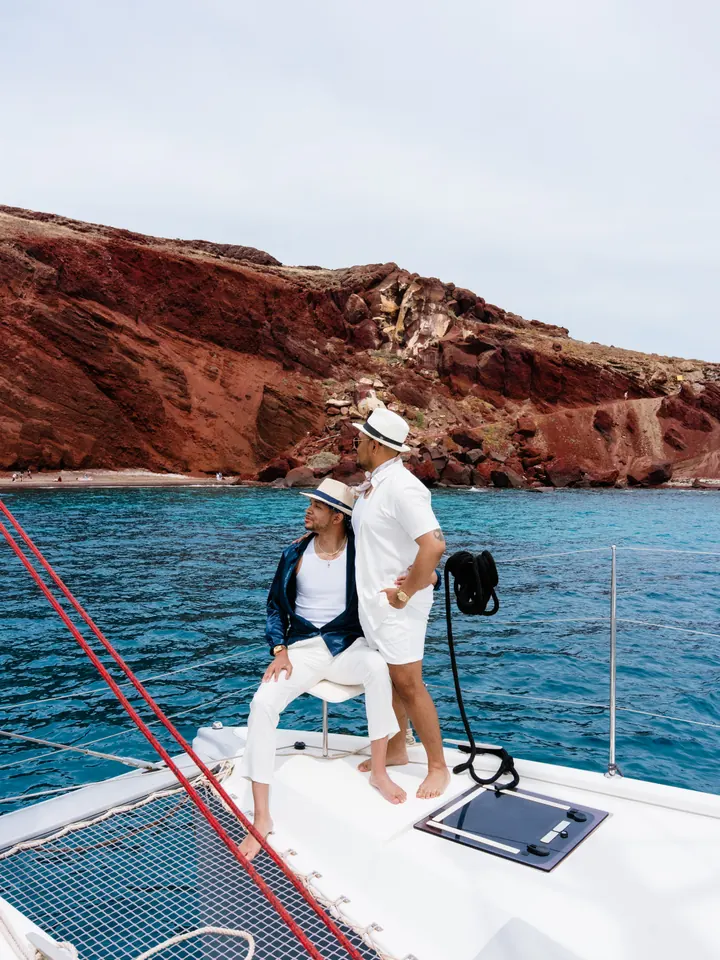 A couple relax on a catamaran bow in front of the dramatic Red Beach cliffs of Santorini