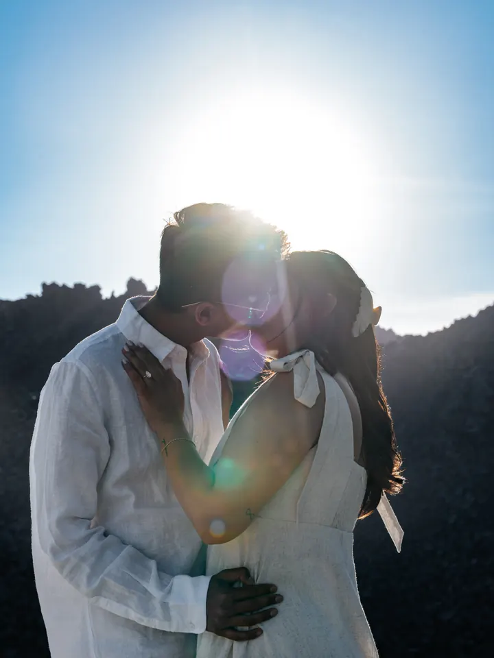 A couple kiss backlit by golden sun flare on the Santorini coast