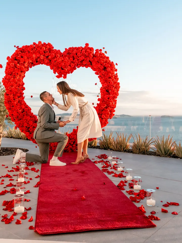 A couple at a red-rose heart arch on a caldera-edge terrace with the blue Aegean behind them