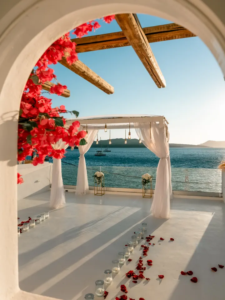 View through a white Cycladic archway draped in pink bougainvillea to a floral proposal arch above the Santorini caldera