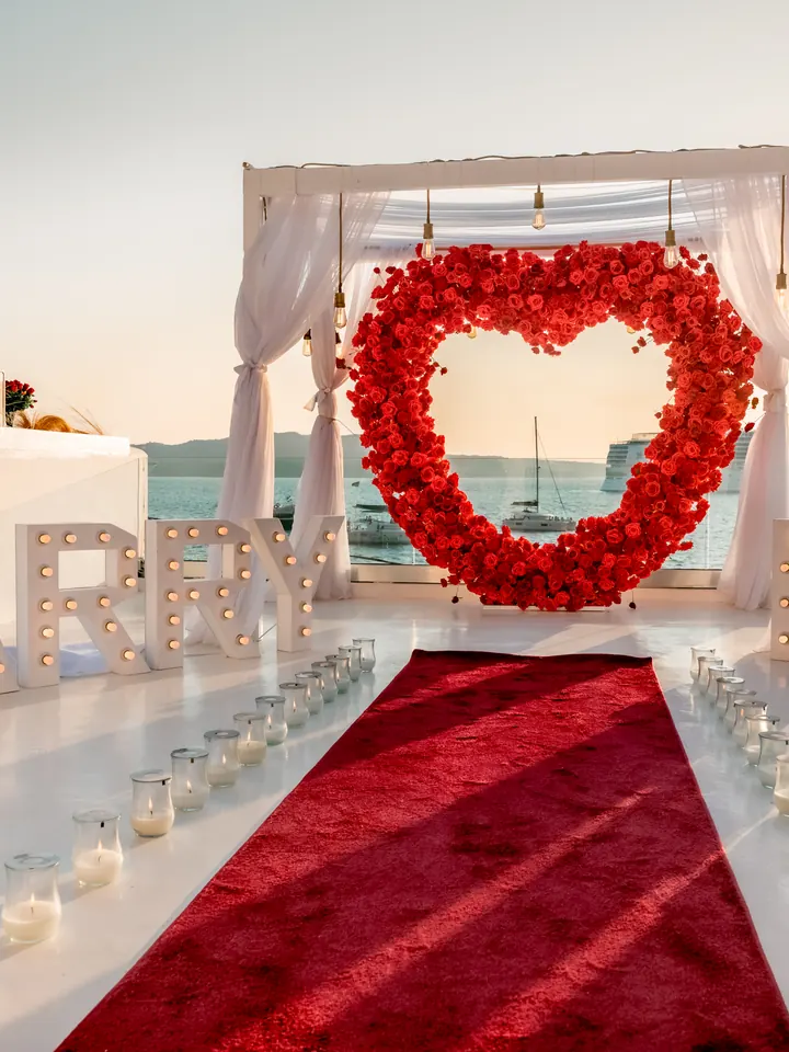 A red-rose heart and MARRY ME letters with a red carpet aisle on an Oia cliff terrace at sunset