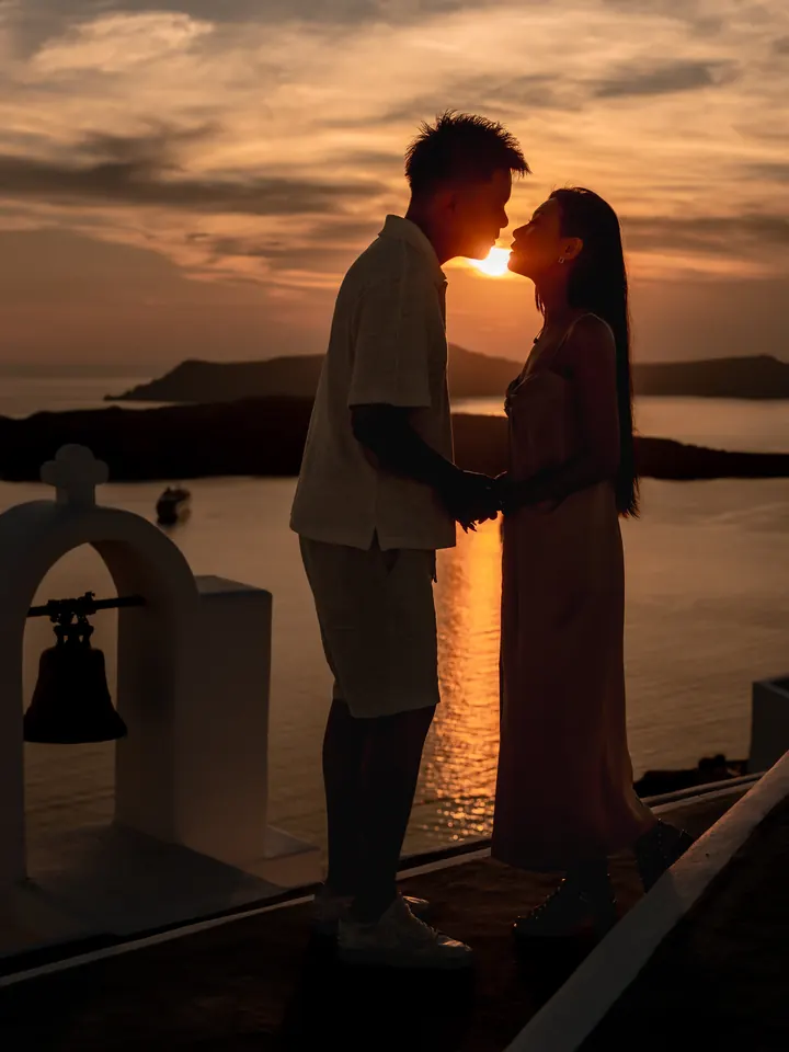 A couple kiss in silhouette against a glowing Santorini sunset beside a white chapel bell