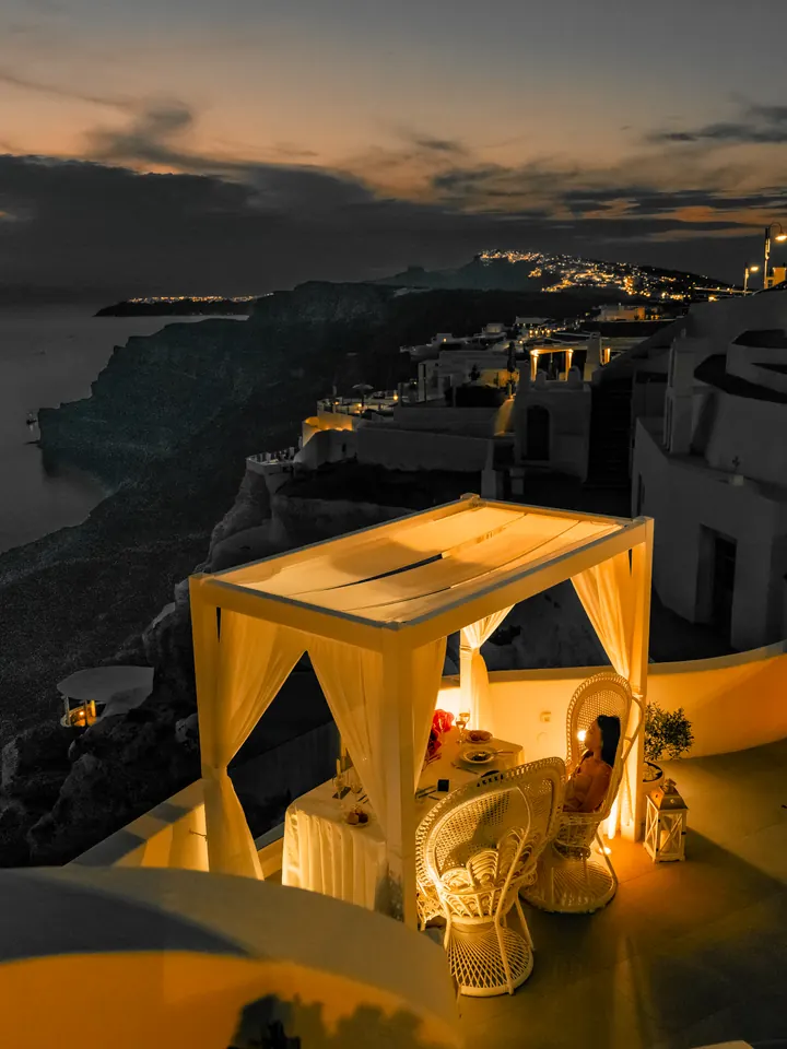 A draped cabana lit with candles on a private terrace as the lights of the Santorini caldera glow at dusk