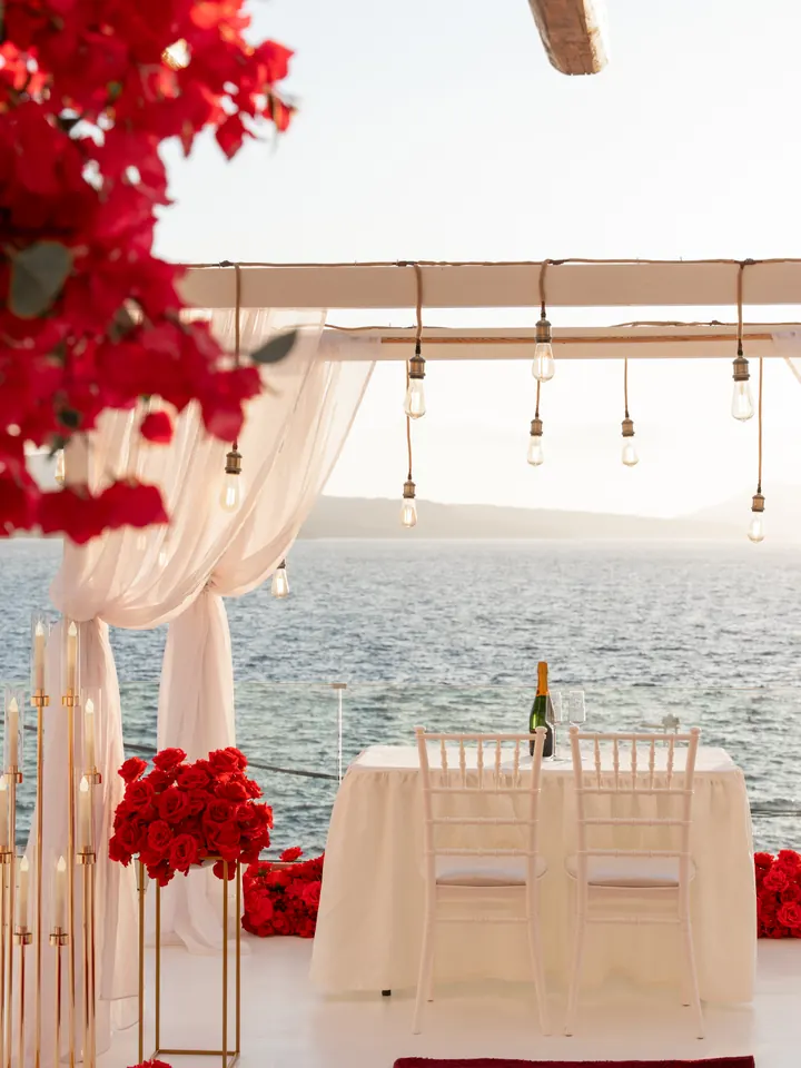 A candlelit private dinner table set with champagne and red roses on a Santorini terrace overlooking the sea