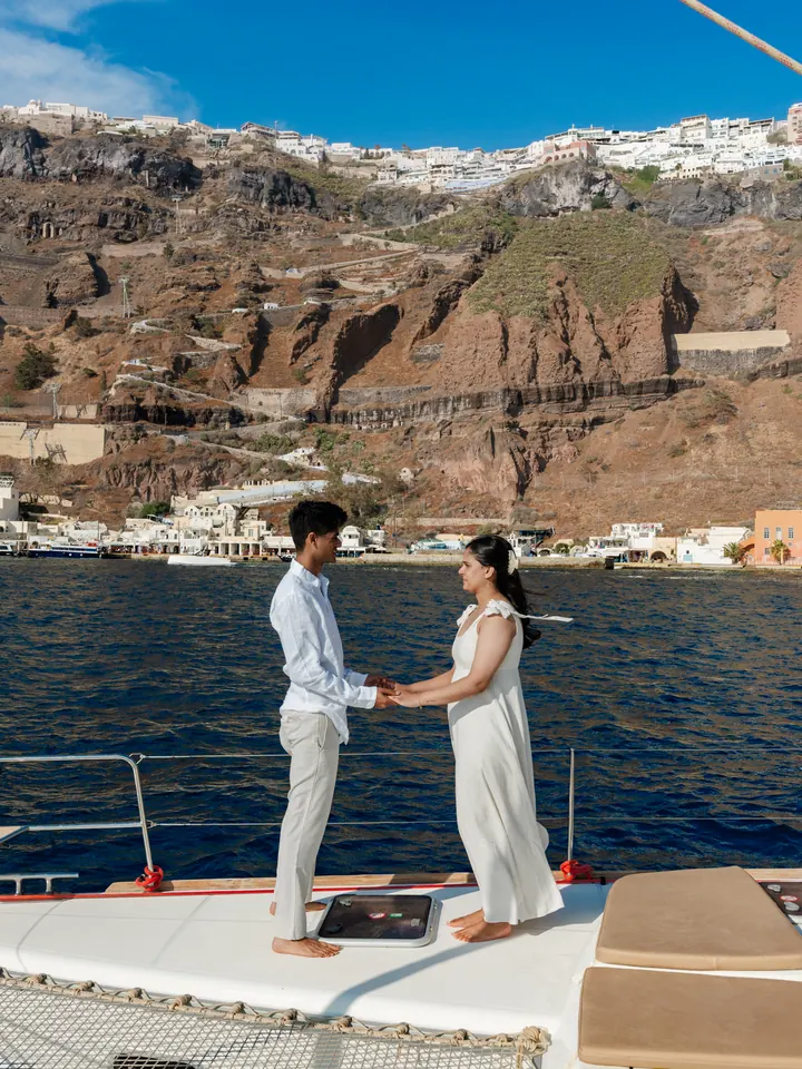 A couple hold hands at the bow of a private catamaran beneath the Santorini cliffs