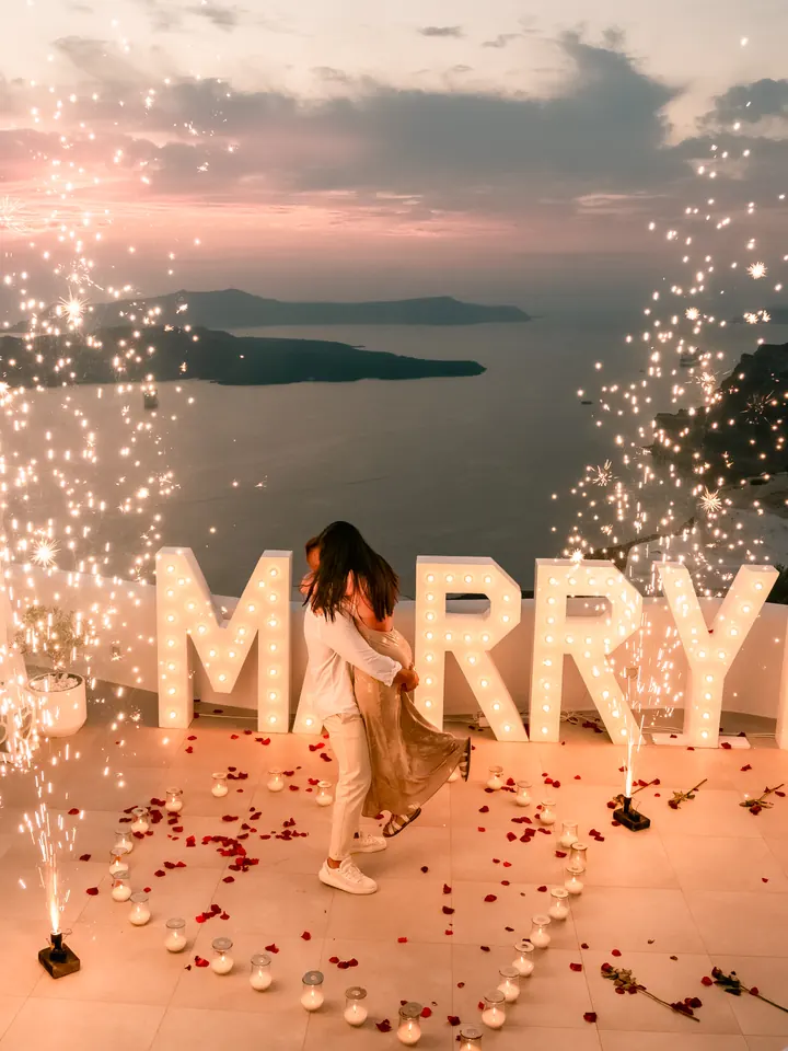 A woman in a flowing dress twirls between sparkling fountains beside glowing MARRY ME letters at a pink Santorini sunset