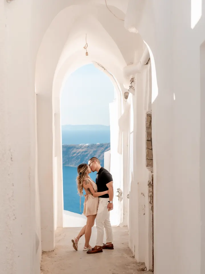 A couple kiss in a sunlit white Cycladic passageway framing the blue Santorini caldera