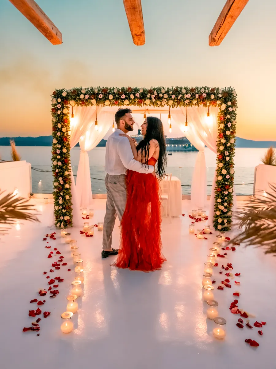 A couple embrace beneath a candlelit floral arch on a Santorini caldera terrace at sunset, the woman in a red gown