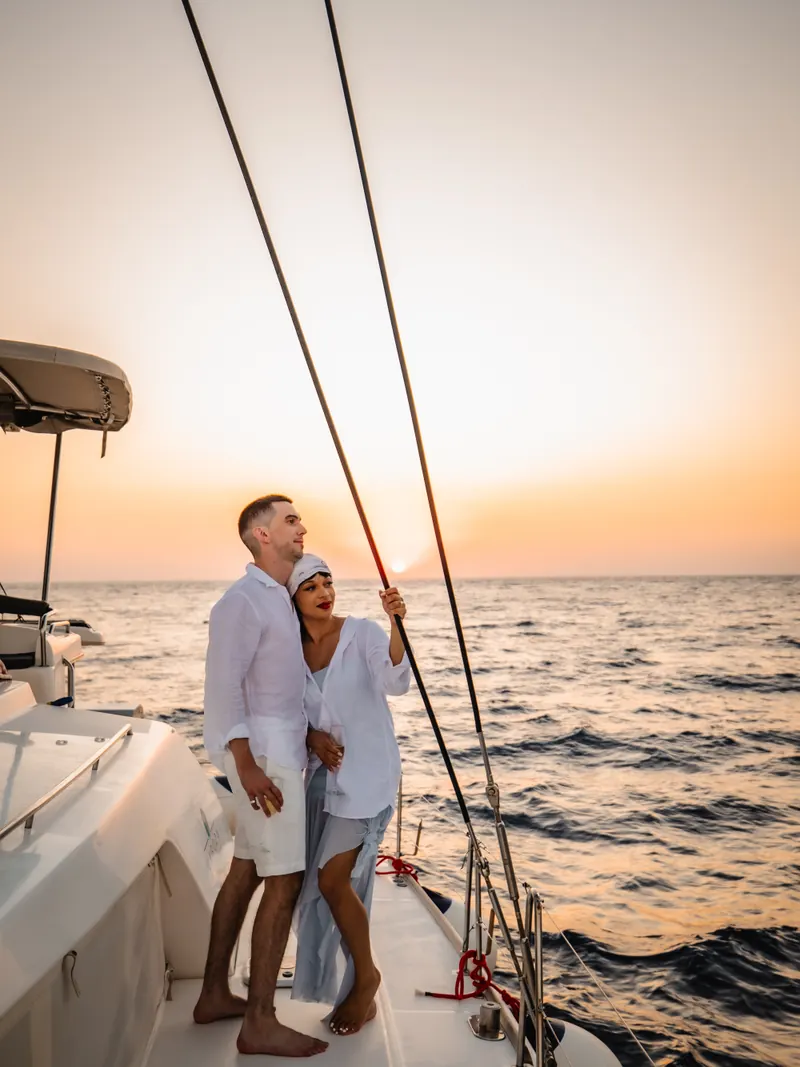 A couple stand at the bow of a private catamaran watching the sun set over the sea near Santorini