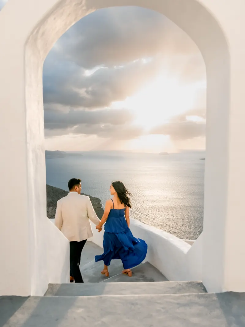 A couple in evening dress walk hand in hand through a white Cycladic archway toward a Santorini sunset
