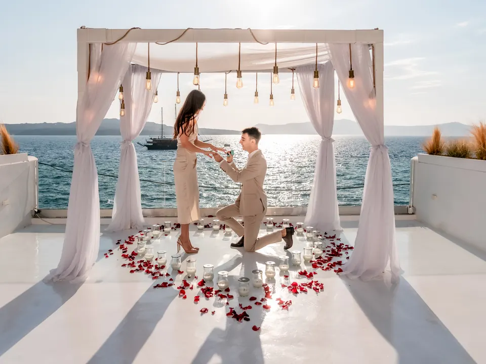 A cliffside proposal setup with a draped white arch and hanging lights overlooking the sunlit Santorini caldera
