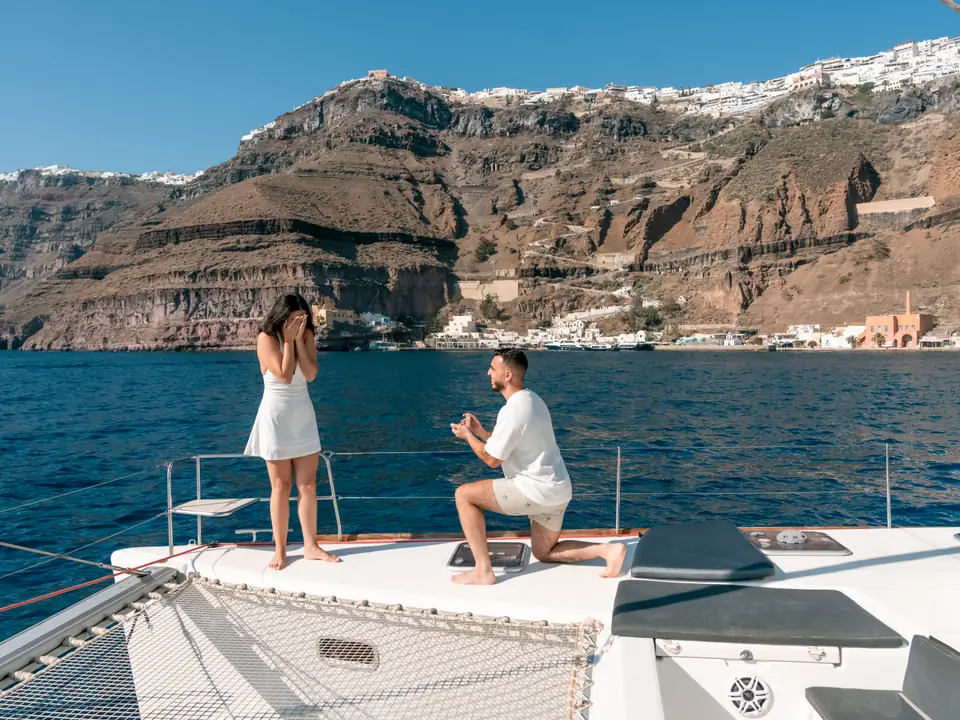 A man kneels to propose on the deck of a private catamaran beneath the Santorini cliffs