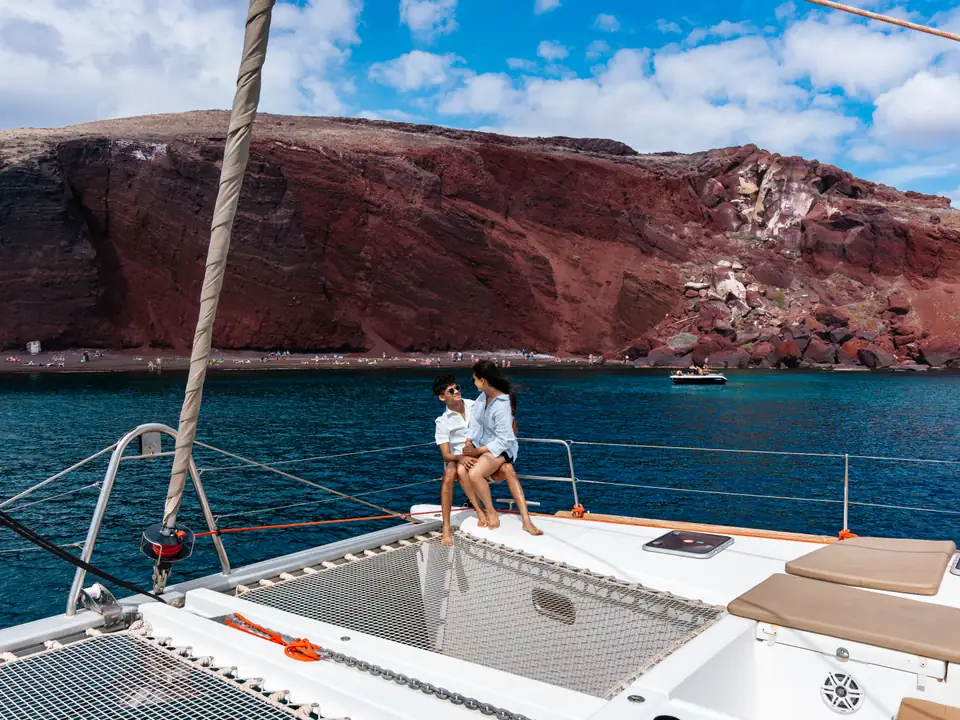 A couple on a catamaran bow in front of the dramatic red volcanic cliffs of Red Beach, Santorini