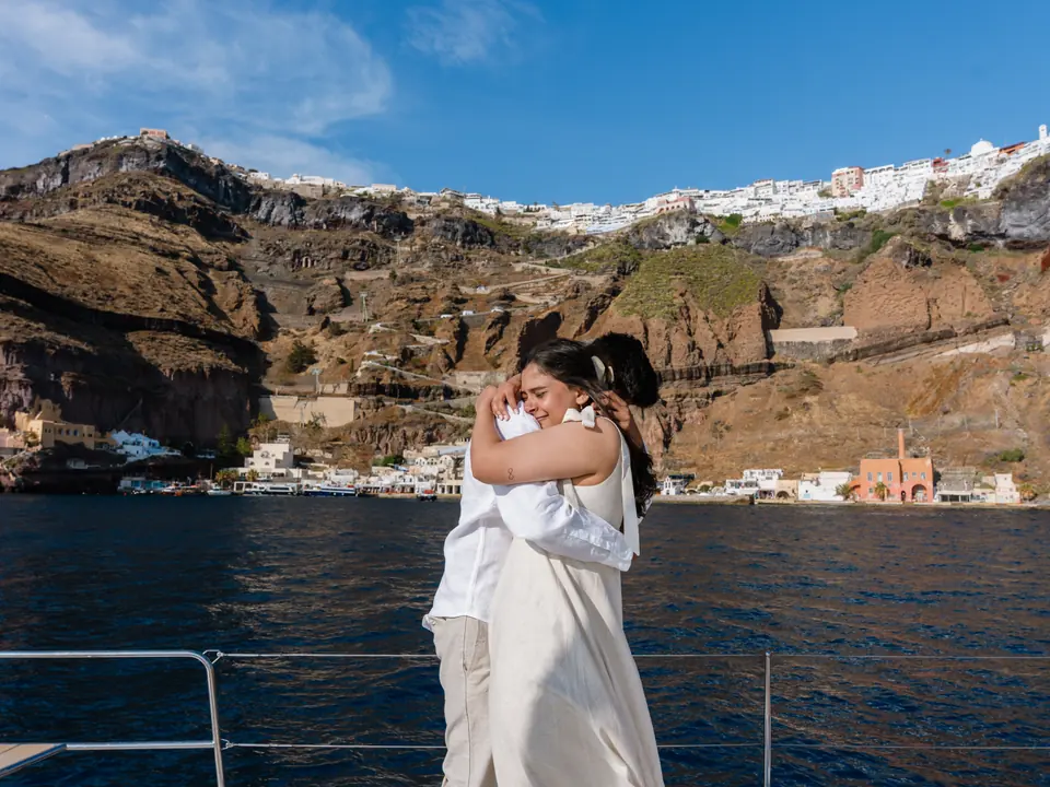 A couple embrace on a catamaran with the cliffside town of Fira rising behind them in Santorini