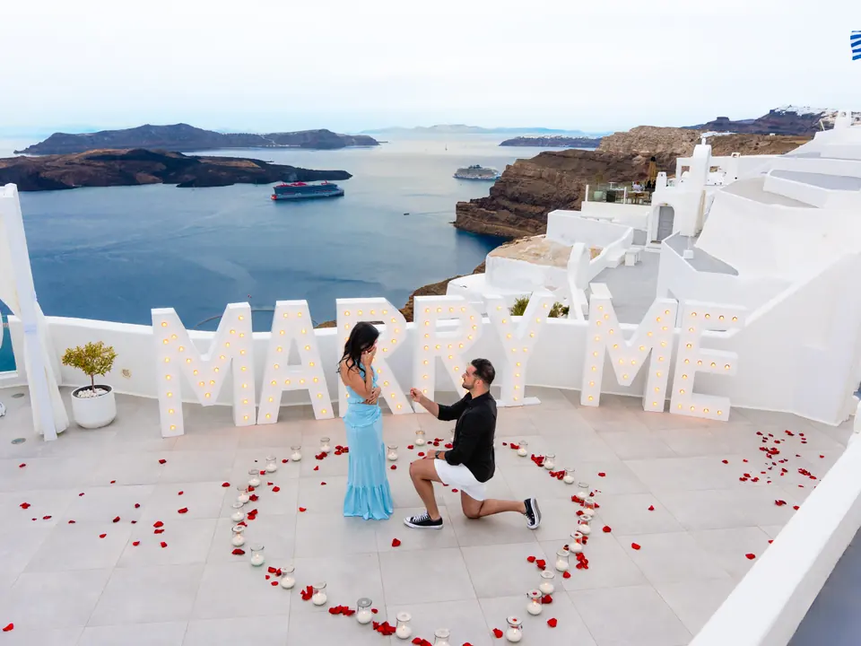 A couple at a MARRY ME proposal setup on a high Imerovigli terrace with the panoramic Santorini caldera behind them