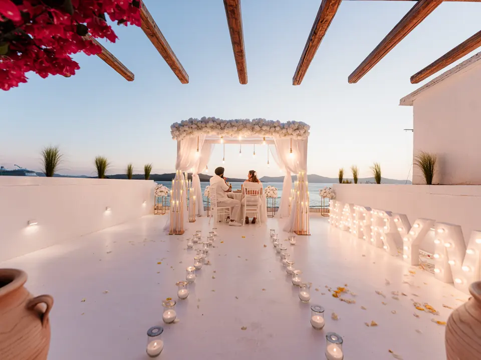 A white-draped proposal setup with glowing MARRY ME letters on an Oia caldera terrace as dusk settles over the sea