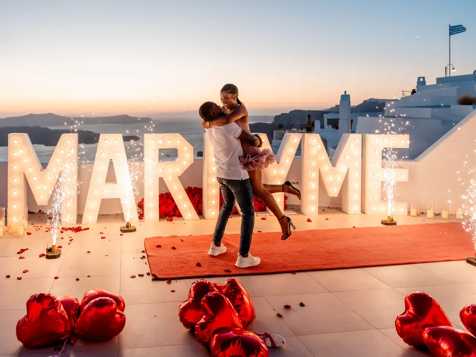 A couple embrace between sparkling fountains and glowing MARRY ME letters with red heart balloons on a Santorini terrace at dusk