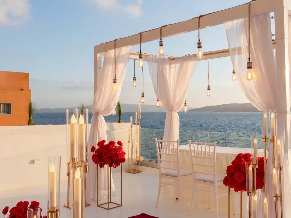 A white floral proposal arch with red roses, gold candelabra and chairs set on a Santorini terrace above the sea