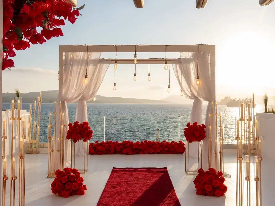 A red carpet aisle lined with roses and candles leading to a draped arch on an Oia cliff terrace at sunset