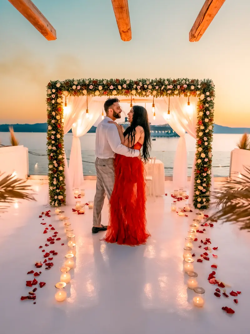 A couple embrace beneath a candlelit floral arch on an Oia cliff terrace at sunset, the woman in a red gown, a rose-petal aisle leading to the sea