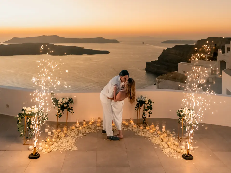 A couple kiss between sparkling fountains on a Santorini cliff terrace as the sun sets over the caldera