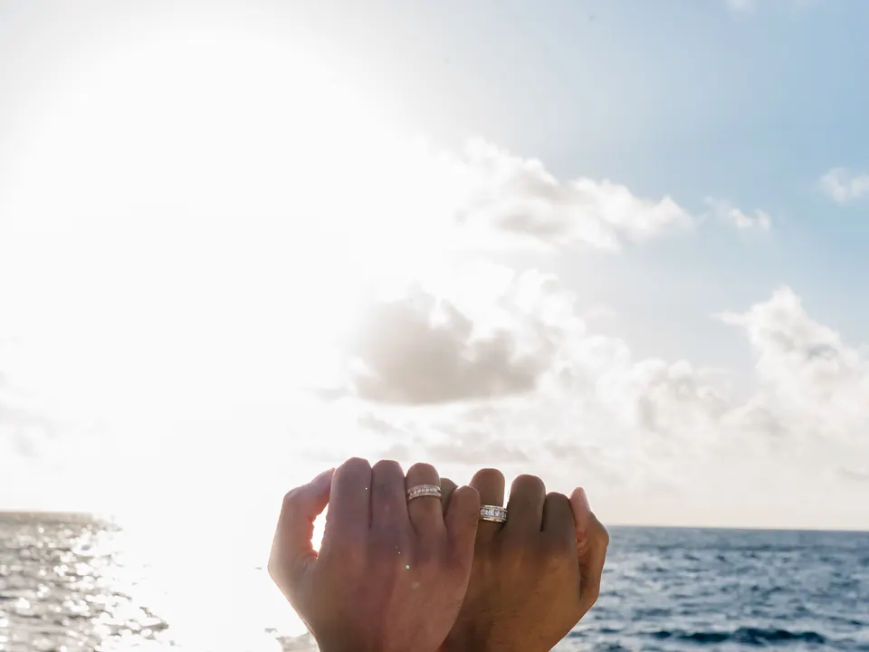 A newly engaged couple raise their hands to show their rings against the bright Aegean sky