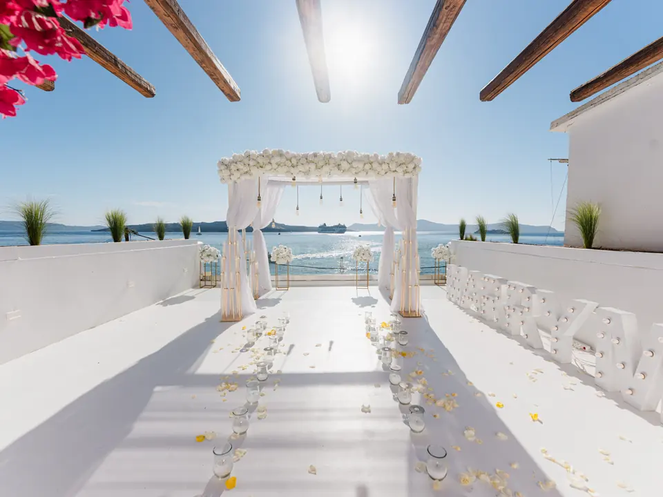 A finished white floral arch and flower-lined aisle styled on a sunlit Santorini caldera terrace