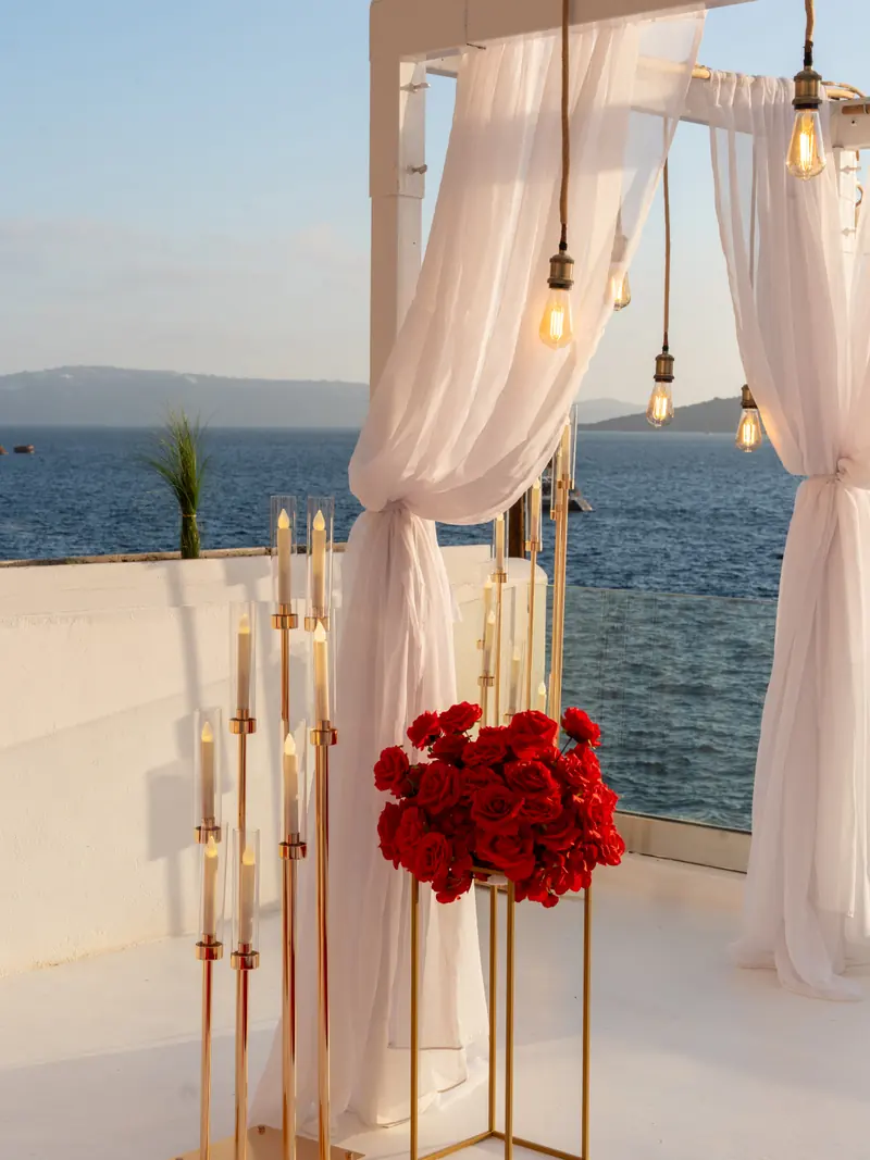 Gold candelabra holding tall candles beside a cluster of red roses and a white-draped arch on a Santorini terrace