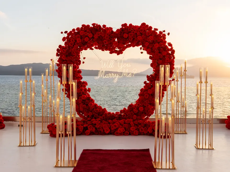 A styled red-rose heart arch with gold candelabra and a red carpet aisle on a Santorini terrace above the sea